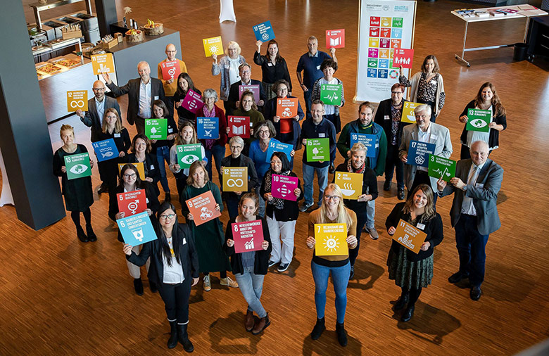 Aus höherer Perspektive sieht man circa 40 Personen, die Schilder mit den globalen Nachhaltigkeitssymbolen in die Kamera halten.